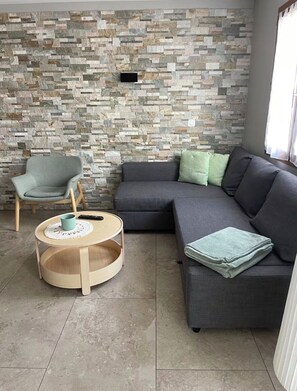 Un salon avec un mur en pierre, un canapé gris, une table basse ronde en bois et un fauteuil bleu clair.