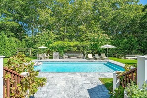 Ein rechteckiger Pool mit einer gefliesten Terrasse, umgeben von üppigem Grün und einem mit Sitzgelegenheiten ausgestatteten Pergola.