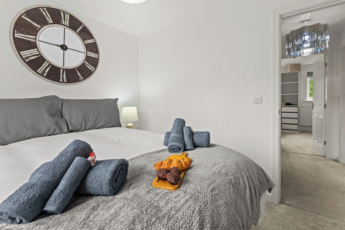 A bedroom with a large wall clock, a bed with grey bedding, rolled towels, and a tray with pastries.