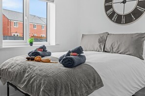 Une chambre à coucher avec un lit, une grande horloge murale, une fenêtre donnant sur des immeubles et une plante en pot posée sur le rebord de la fenêtre.