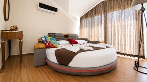 A round bed with a headboard, a bedside table, a lamp, and a window with curtains.