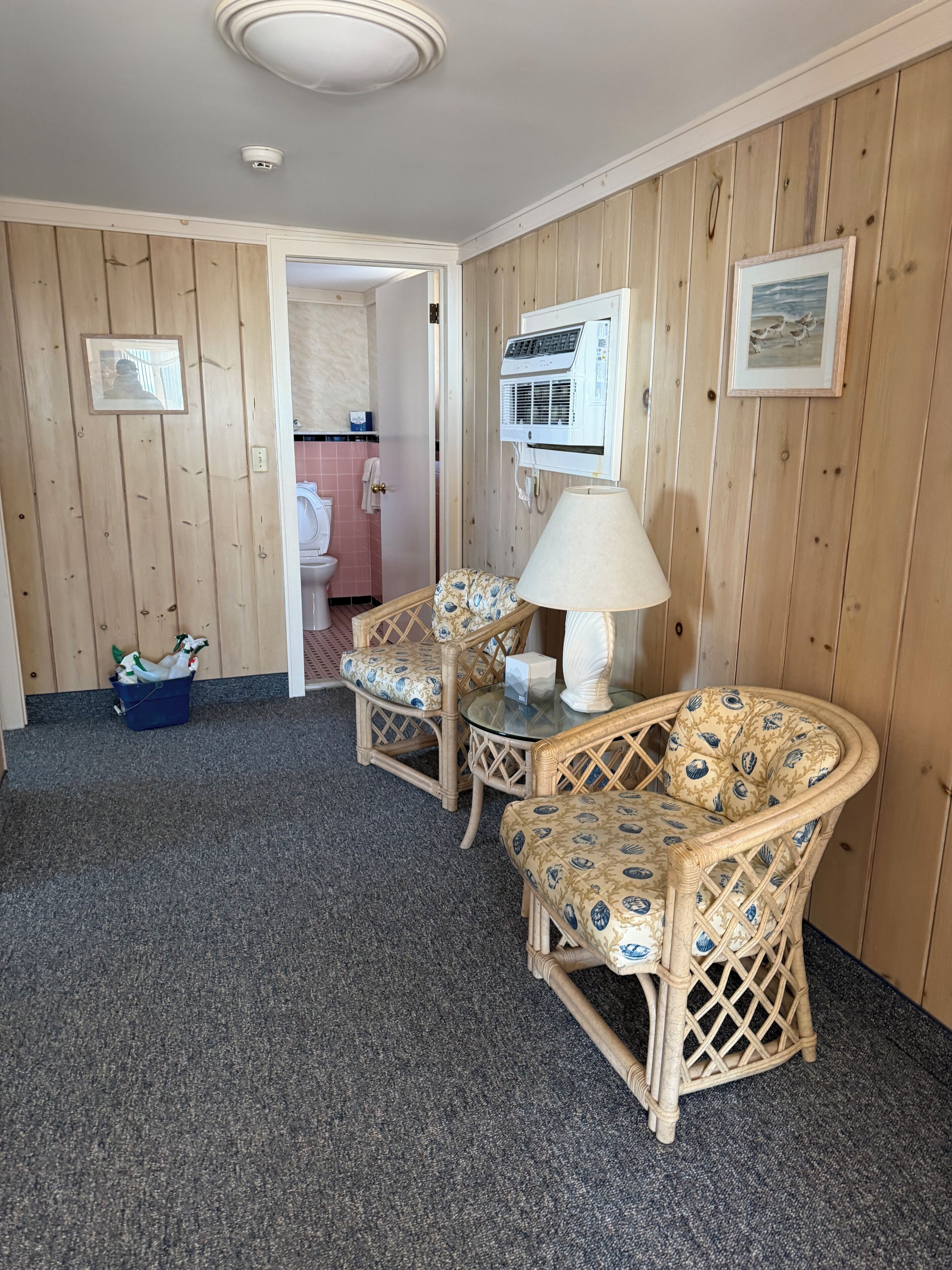 Ein Zimmer mit Holzwandverkleidung, ein Korbstuhl mit Kissen, ein kleiner Tisch mit Lampe und ein blauer Korb auf dem Boden.