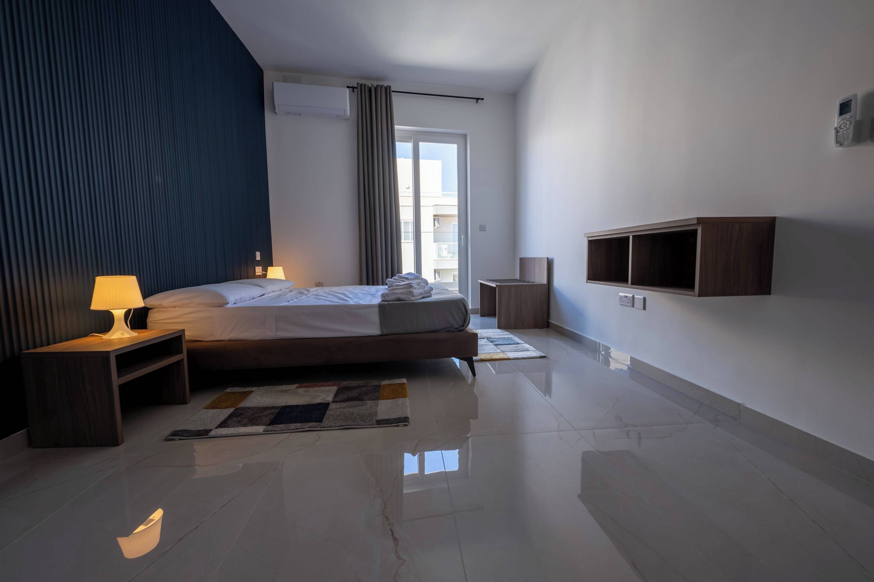 Een moderne hotelkamer met een bed, bedlampen, een klein houten nachtkastje, een ingebouwde tv-kast, een groot raam met gordijnen en een betegelde vloer.
