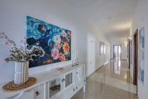 A hallway with a large floral painting on the wall, a white console table with decorative items, and a potted plant. - Ghajnsielem 3BDR Flat with Views - NO Lift (Għajnsielem)