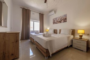 A hotel room with a bed, bedside tables, a wooden dresser, a window with curtains, and a wall-mounted air conditioner. - Ghajnsielem 3BDR Flat with Views - NO Lift (Għajnsielem)