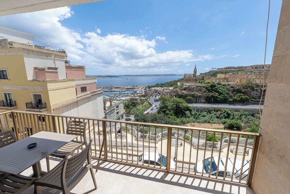 A balcony with a table and chairs, overlooking a coastal town with buildings and a church. - Ghajnsielem 3BDR Flat with Views - NO Lift (Għajnsielem)
