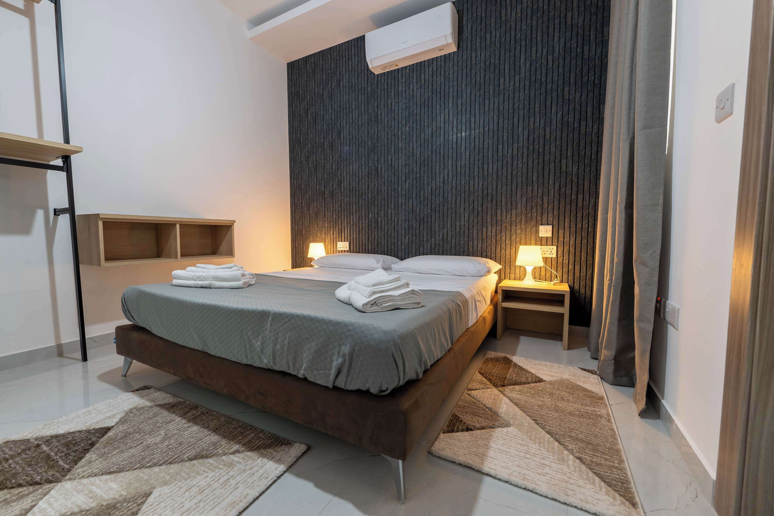 A modern bedroom with a bed, bedside lamps, a wooden headboard, a wall-mounted air conditioner, and a built-in shelf.