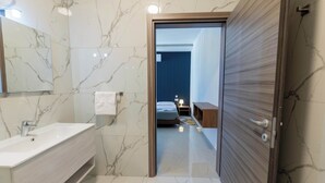 Een moderne badkamer met een marmeren vloer, een wit wastafelmeubel en een houten deur die leidt naar een slaapkamer met een bed en nachtkastje.