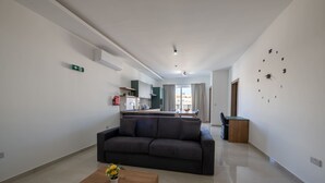 Uma sala de estar moderna com sofá, mesa de centro de madeira com flores, cozinha ao fundo e um relógio de parede grande.