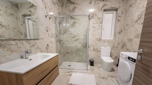 Um banheiro com piso de mármore, toucador de madeira, box de vidro, vaso sanitário e máquina de lavar.