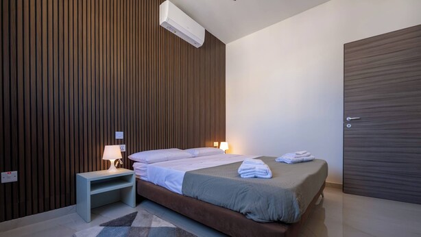 Quarto moderno com cama, mesa de cabeceira, abajur e ar-condicionado fixado na parede.