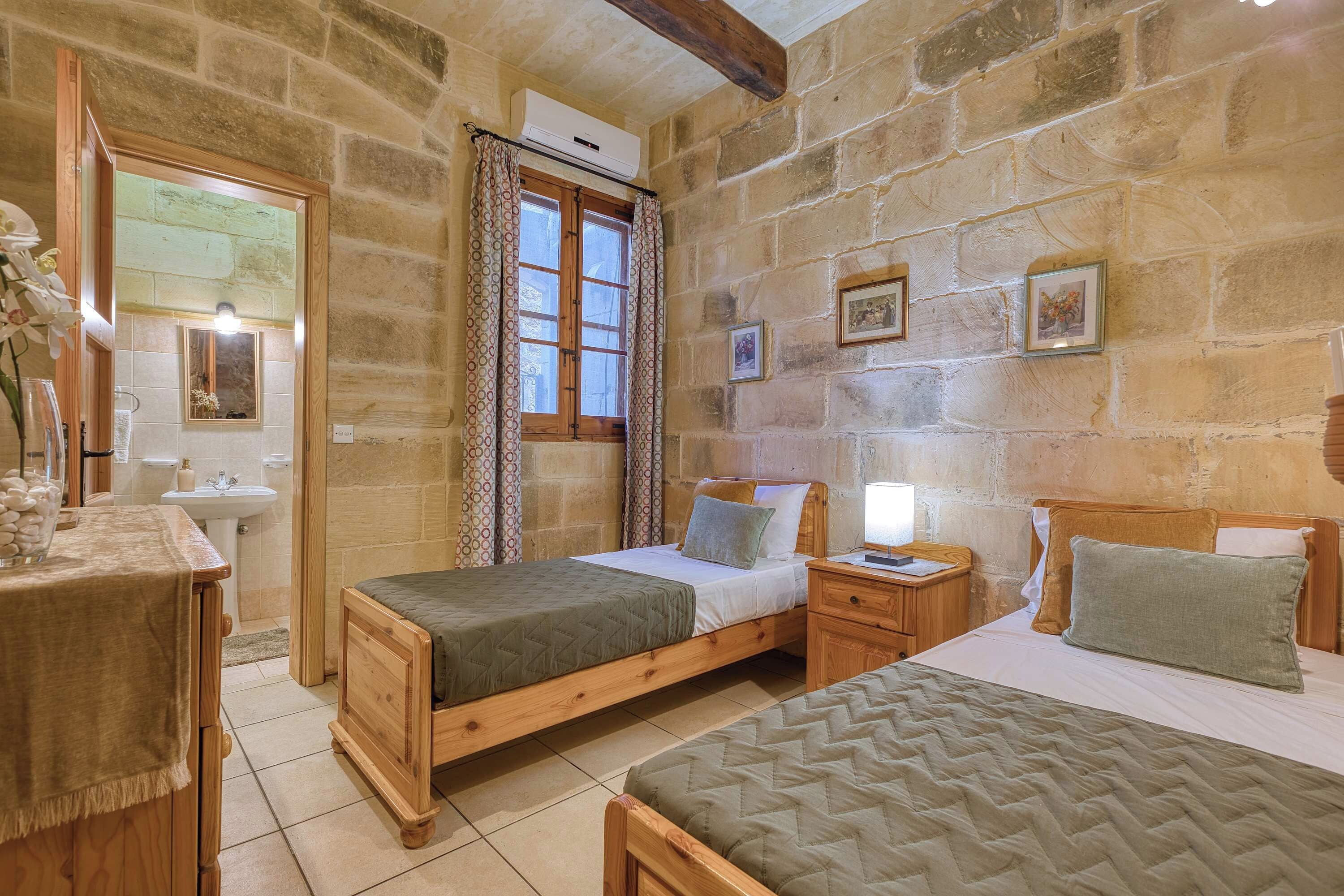 Una habitación con paredes de piedra, una cama de madera, una mesita de noche con una lámpara, un espejo y una ventana con cortinas estampadas.
