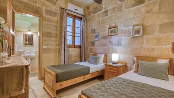 Una habitación con paredes de piedra, una cama de madera, una mesita de noche con una lámpara, un espejo y una ventana con cortinas estampadas.
