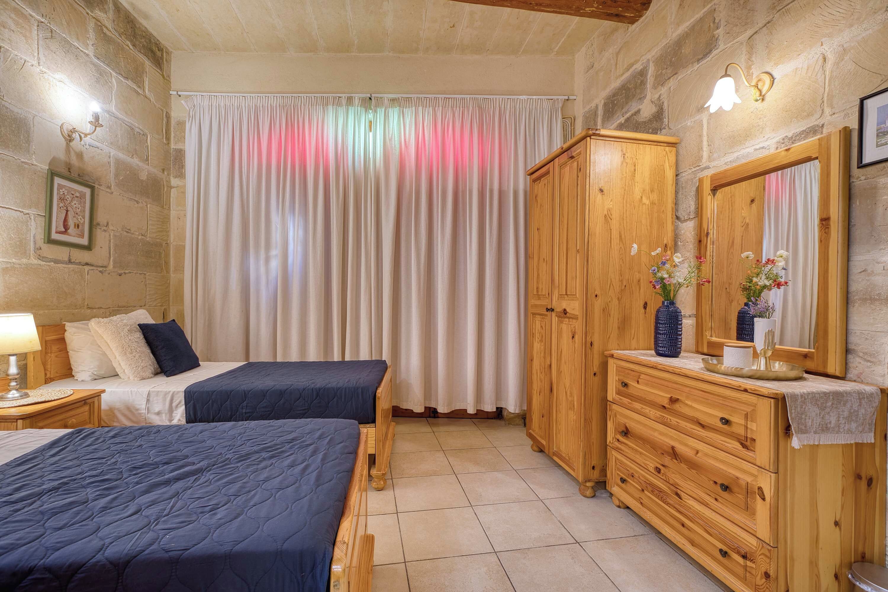 A bedroom with two beds, a wooden wardrobe, a dresser, a mirror, and a nightstand with a lamp.