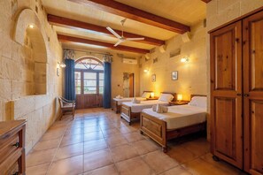 A room with two beds, a wooden wardrobe, a ceiling fan, and stone walls.