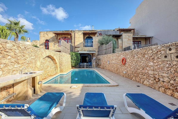 A pool area with lounge chairs, a stone wall, and a building with arched windows.