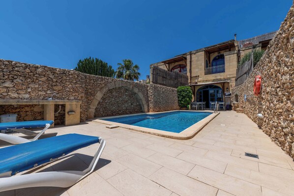 A pool area with stone walls, lounge chairs, and a building with balconies.