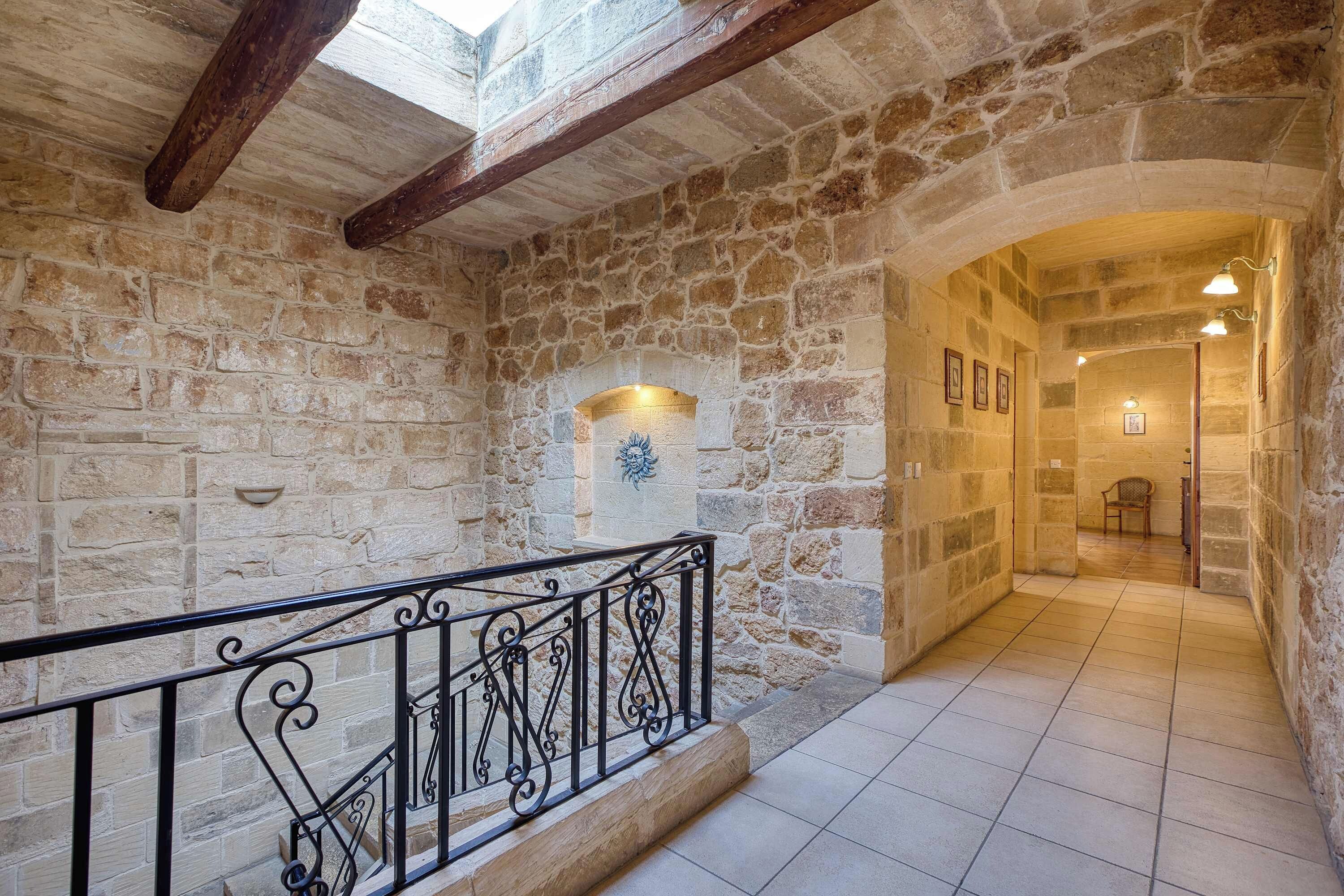 Interior con paredes de piedra, una escalera que lleva a un nivel superior, suelo de baldosas y un acceso a otra habitación.