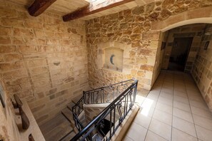 Un intérieur aux murs de pierre, avec un escalier, une petite alcôve dotée d’une fenêtre en forme de lune et un sol carrelé.