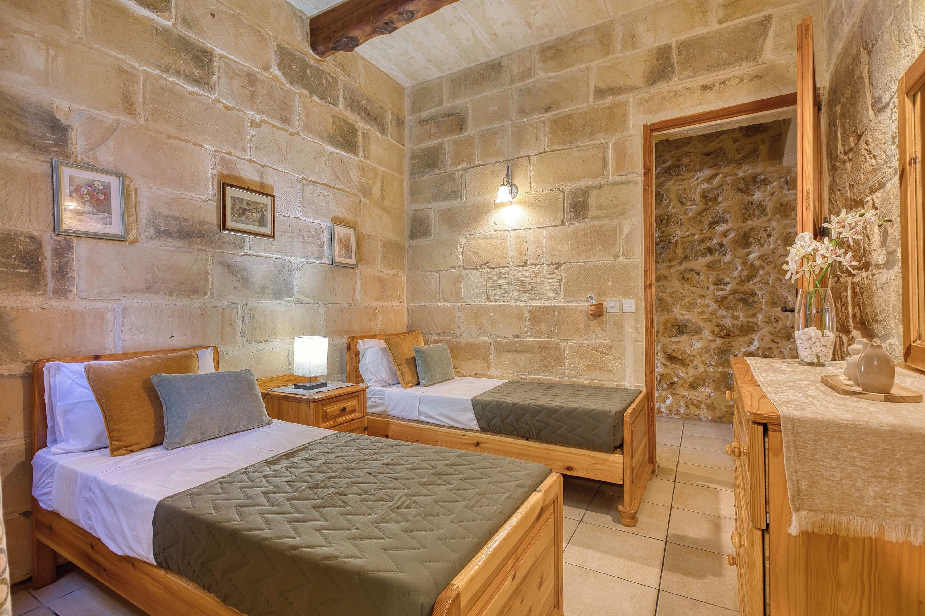 Una habitación con dos camas de madera, una pared de piedra, una mesita de noche de madera con una lámpara y una pequeña mesa con un jarrón de flores.