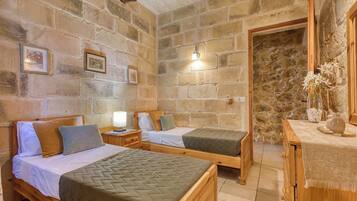 Una habitación con dos camas de madera, una pared de piedra, una mesita de noche de madera con una lámpara y una pequeña mesa con un jarrón de flores.