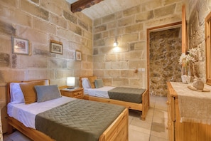 A room with two wooden beds, a stone wall, a wooden nightstand with a lamp, and a small table with a vase of flowers.