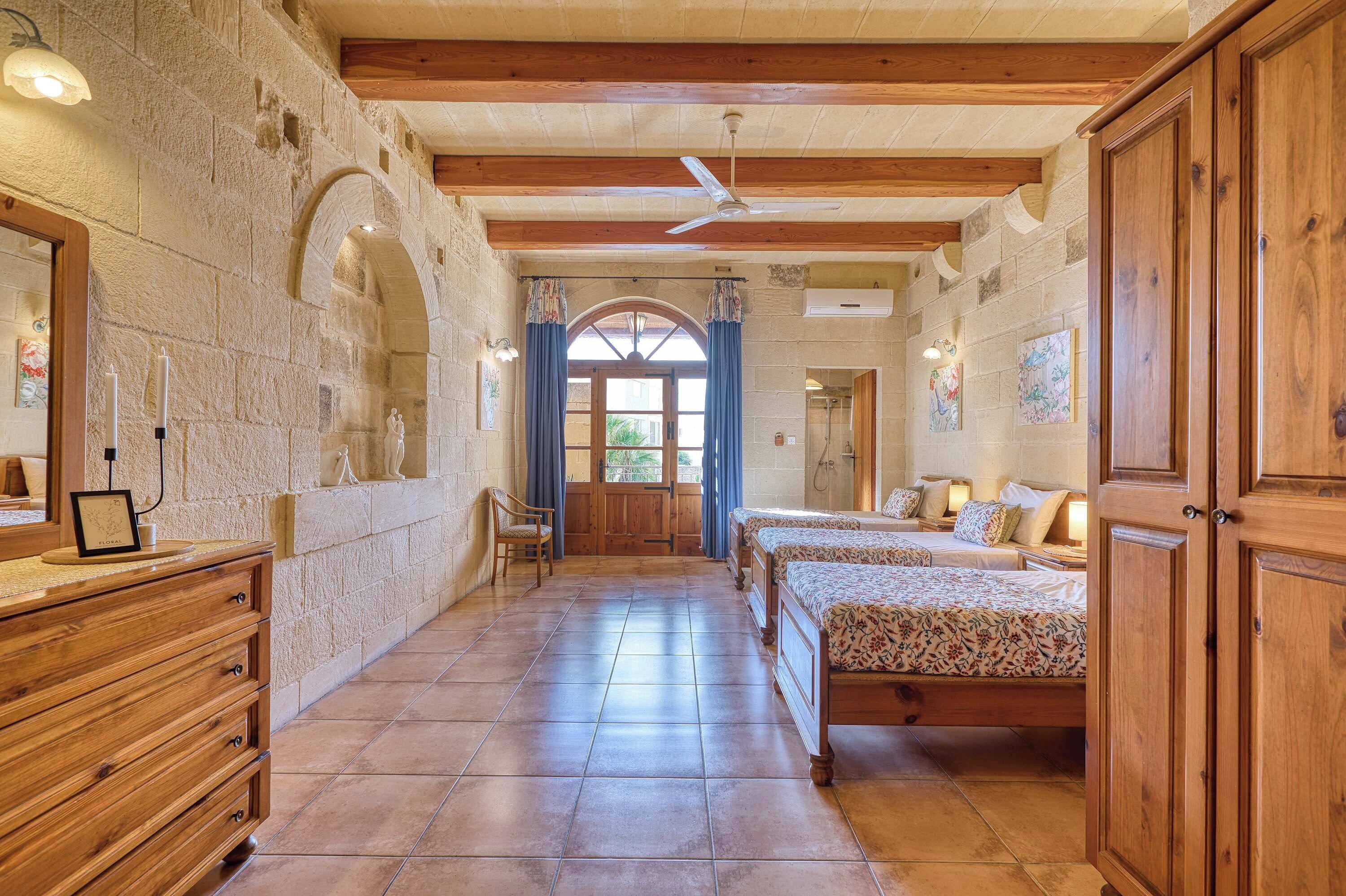 Habitación de hotel con pared de piedra, vigas de madera, cama con ropa de cama floral, cómoda de madera y zona de ducha.