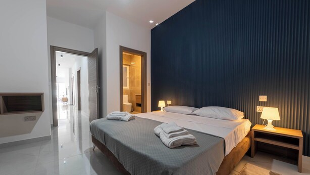 Ein modernes Hotelzimmer mit einem großen Bett, Nachttischlampen, einem Holfnachtkästchen und Blick auf einen Flur.