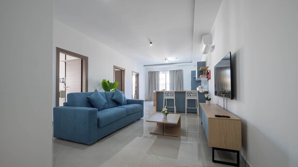 A modern living room with a blue sofa, a coffee table, a TV, and a dining area. - Swieqi Brand New 3BDR Flat- Spacious Getaway - FL7 (Is-Swieqi)