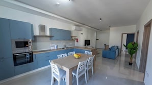 A modern kitchen with a central island, built-in appliances, and a dining area with white chairs. - Swieqi Brand New 3BDR Flat- Spacious Getaway - FL7 (Is-Swieqi)