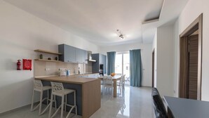 A modern kitchen with a central island, white bar stools, and a dining area with a table and chairs. - Stunning New 3BDR Apartment in Swieqi - FL9 (Is-Swieqi)