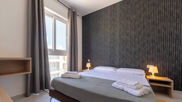 Une chambre d’hôtel moderne avec un grand lit, deux lampes de chevet, une table de nuit en bois, une fenêtre avec des rideaux et une vue sur les bâtiments environnants.