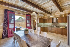 A rustic kitchen with wooden cabinets, a dining area with a table and chairs, and a red door with glass panels. - Tan Nanna 5 BDR Farmhouse by PMI Malta (Ix-Xagħra)
