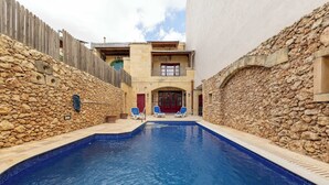 Ein Schwimmbad mit Steinmauern und einem Holzzaun, zwei Liegestühle und ein Gebäude mit einem Balkon.