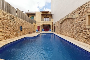 Ein Schwimmbad mit Steinmauern und einem Holzzaun, zwei Liegestühle und ein Gebäude mit einem Balkon.