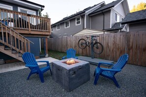 Ein Garten mit einem Feuerkorb, zwei Adirondack-Stühlen, einer Holzterrasse und einem Fahrrad.