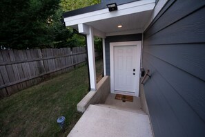 Une maison avec une porte blanche, un petit porche et une clôture en bois à l’arrière-plan.