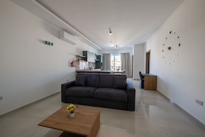 Ein modernes Wohnzimmer mit einer Couch, einem Couchtisch mit Blumen, einer Küche im Hintergrund und einer großen Wanduhr.