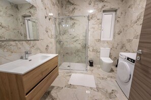 Ein Badezimmer mit Marmorboden, Waschtisch aus Holz, Duschkabine aus Glas, Toilette und Waschmaschine.