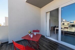 Ein Balkon mit rotem Tisch und Stühlen, einer Topfpflanze und Blick auf Gebäude durch eine Glastür.