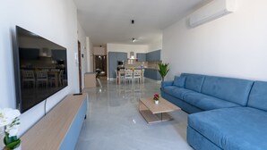 Un salon moderne avec un téléviseur à écran plat, un canapé bleu, une table basse et un coin repas à l’arrière-plan.