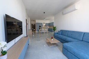 Ein modernes Wohnzimmer mit einem Flachbildfernseher, einem blauen Sofa, einem Couchtisch und einem Essbereich im Hintergrund.