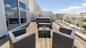 Une terrasse sur le toit, dotée de mobilier en osier, d’un jacuzzi et d’une balustrade en verre offrant une vue sur le paysage urbain.