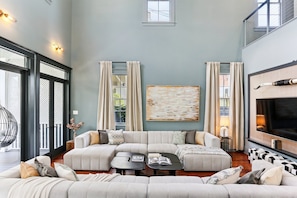 A spacious living room with a large sectional sofa, a coffee table, and a fireplace. There are curtains, a painting, and a large window providing natural light. - Grand 10BR NOLA Retreat, Dual Heated Pools & Spas (New Orleans)