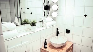 Et moderne badeværelse med hvide fliser, en trækommode, en rund vask, et spejl med lys og en potteplante.