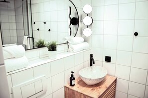 A modern bathroom with white tiles, a wooden vanity, a round sink, a mirror with lights, and a potted plant. - Central Kempten | Sleeps 2 • Stylish (Kempten (Allgäu))