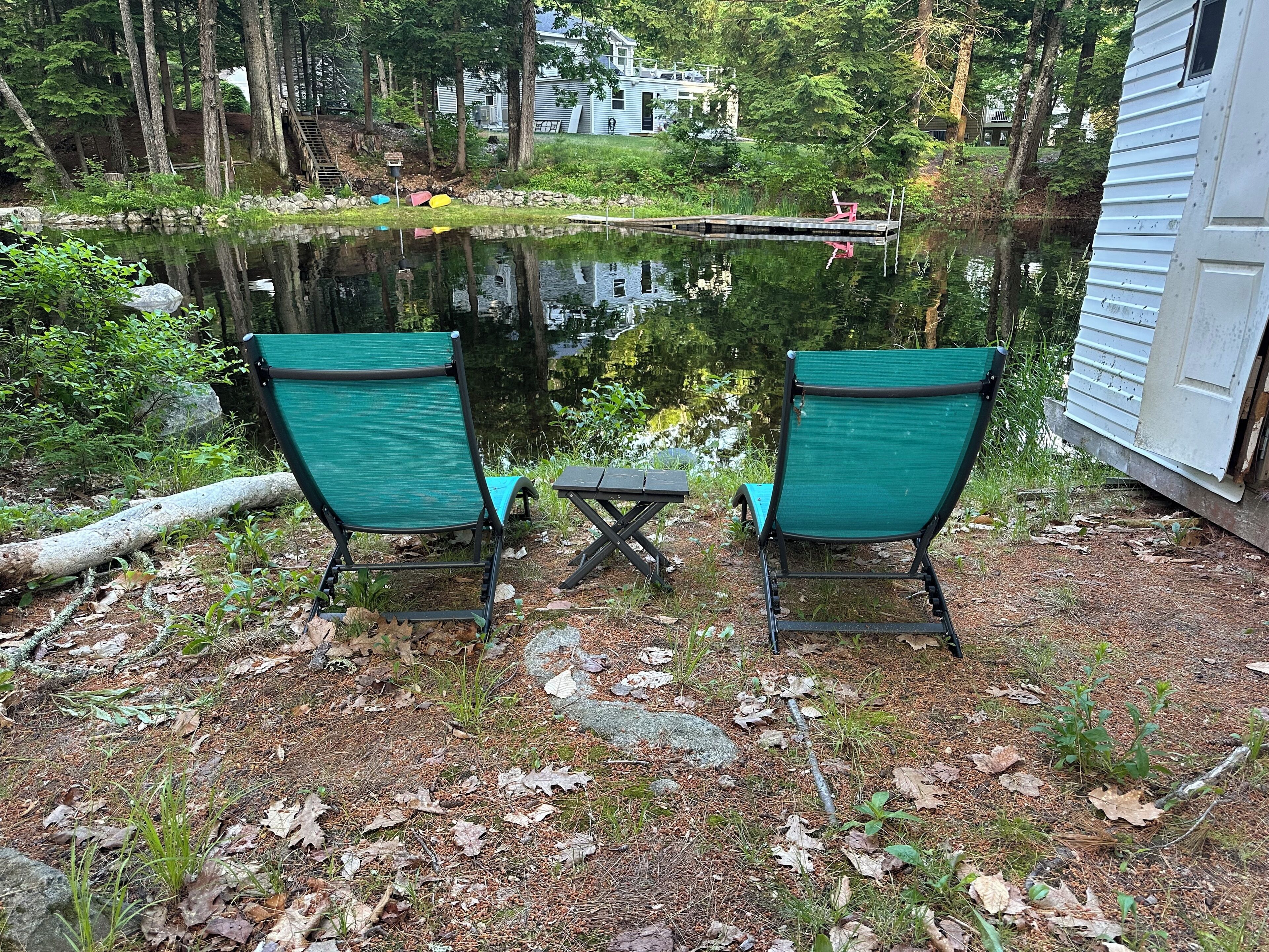 Deux chaises vertes sont placées face à une petite table, près d’un étang doté d’un quai, le tout étant entouré d’arbres et d’une maison.