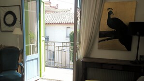 An open door leading to a balcony with a view of buildings and a tiled roof. A chair is visible inside the room, and a painting of a bird is on the wall. - Home 10 minutes from the beaches, between sea and mountains—downtown + garages (Perpignan)