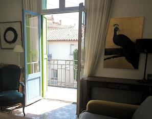 Eine offene Tür führt zu einem Balkon mit Blick auf Gebäude und ein Ziegeldach. Im Raum ist ein Stuhl zu sehen, und an der Wand hängt ein Vogelgemälde.