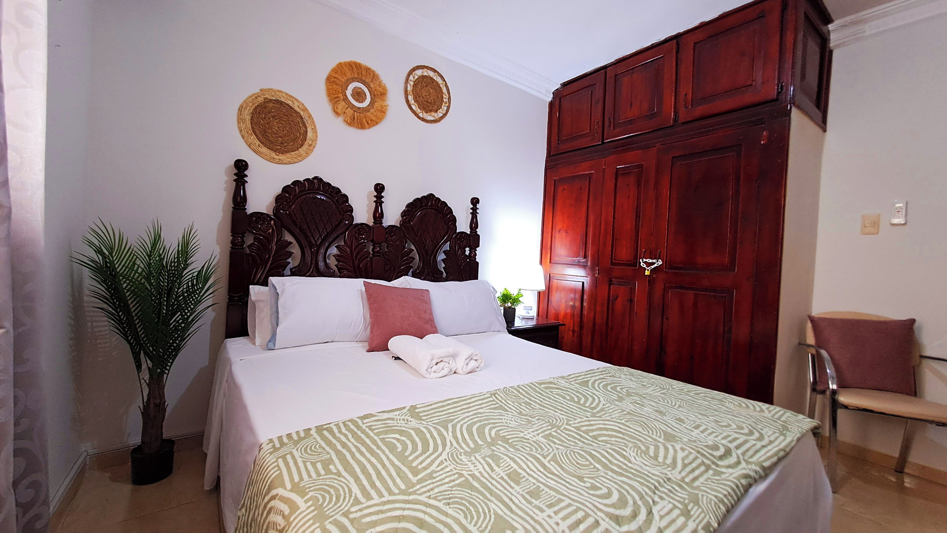 A bedroom with a bed, a wooden wardrobe, a potted plant, and a chair.